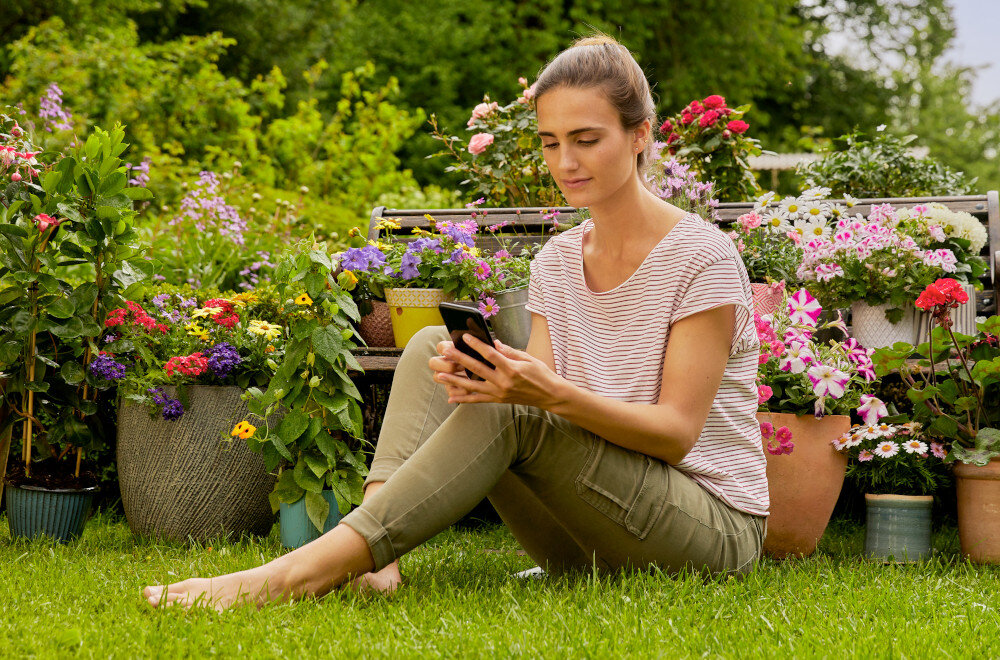 Osoba siedzi na trawie w ogrodzie, trzymając w rękach smartfon. W tle widoczne są różnorodne kwiaty w doniczkach.