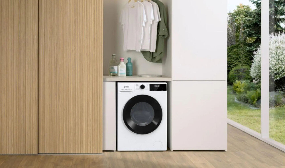 Pralka w zabudowie z widocznym logo \'gorenje\', obok której znajdują się butelki z detergentami i wiszące ubrania. W tle widać ogród za dużym oknem.