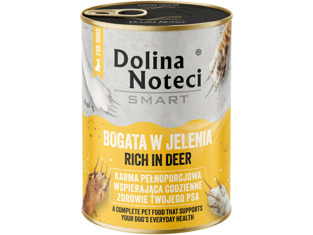 Na podłodze w kuchni stoją dwie puszki karmy dla psa z napisem \'Dolina Noteci Smart Bogata w jelenia\'. Obok puszek siedzi pies, a w tle widać drewniane szafki i miskę na podłodze.