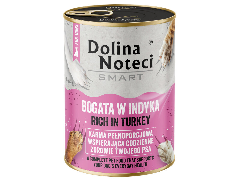 Na drewnianej podłodze stoją trzy puszki karmy dla psa \'Dolina Noteci Smart Bogata w indyka\'. Obok puszek siedzi pies, a w tle widać kuchenne szafki.