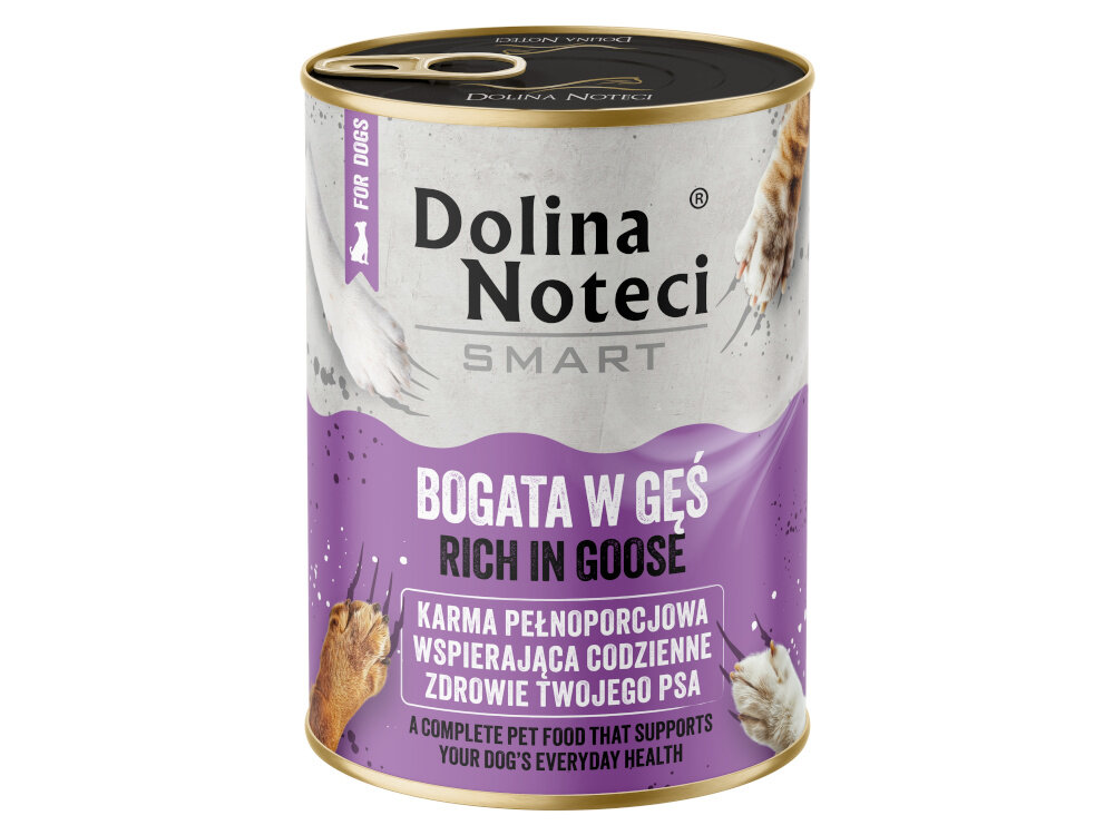 Na podłodze stoi pięć puszek karmy dla psa z napisem \'Dolina Noteci SMART BOGATA W GĘŚ RICH IN GOOSE\'. Obok puszek siedzi pies, a w tle widać kuchenne szafki z produktami.