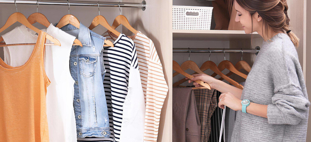 Osoba przegląda ubrania wiszące na wieszakach w szafie, w tym koszulki i kurtkę jeansową. Na półce znajduje się biały koszyk z otworami.