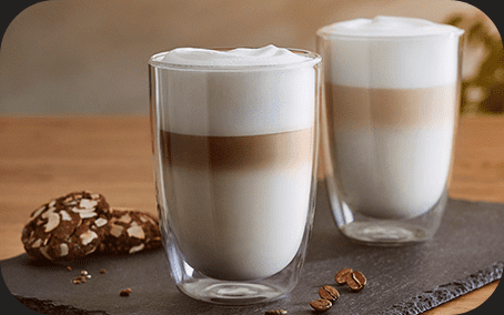Na obrazie znajdują się dwa szklane kubki z napojem warstwowym, prawdopodobnie kawą latte, ustawione na ciemnej tacy. Obok kubków leżą ziarna kawy i ciastka.