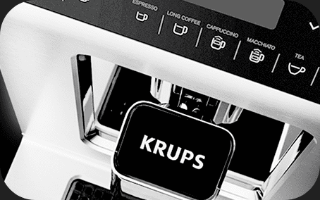 Czarno-biały ekspres do kawy z widocznym logo \'KRUPS\' na przodzie. Na panelu sterowania znajdują się ikony i napisy: ESPRESSO, LONG COFFEE, CAPPUCCINO, MACCHIATO, TEA.