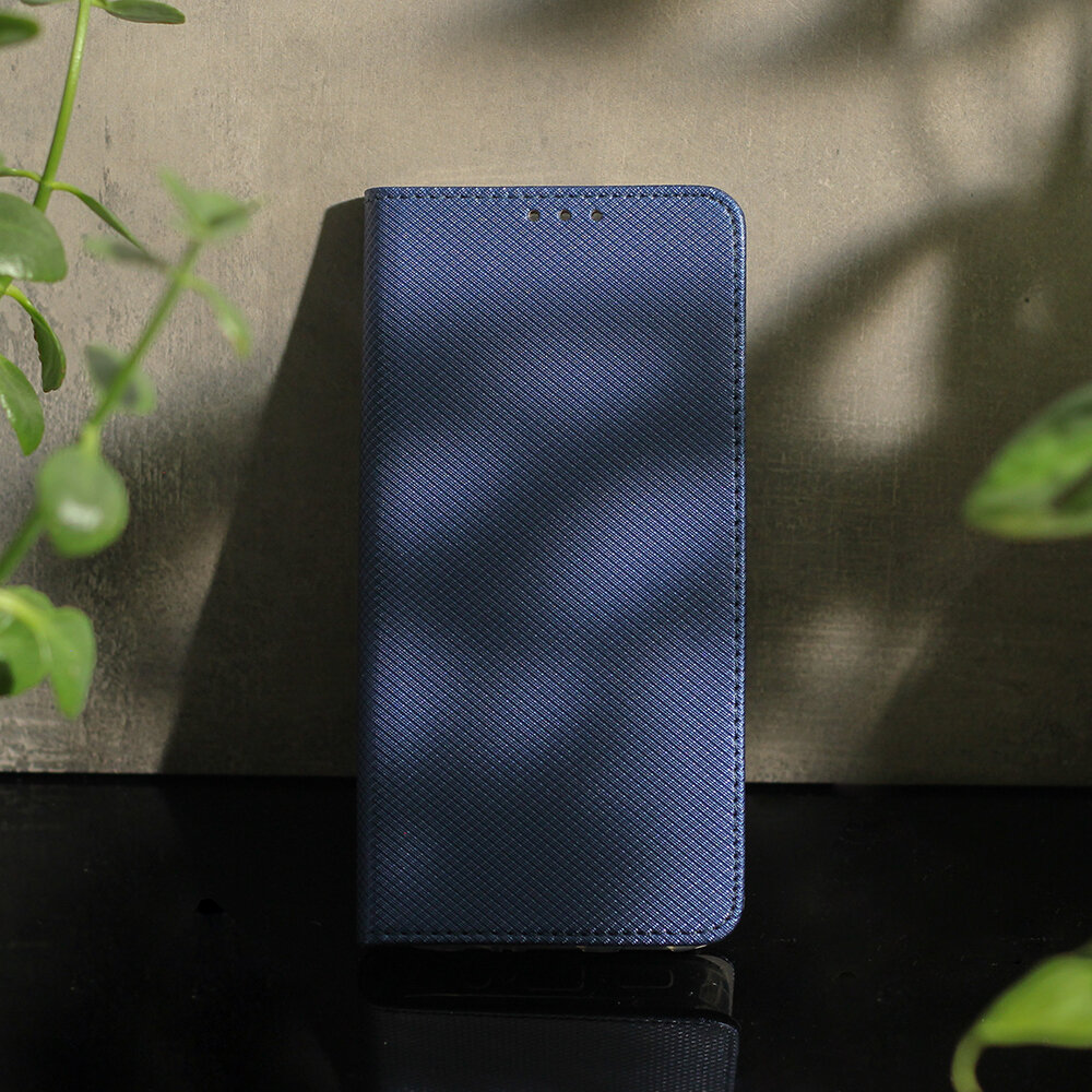 Granatowe etui na telefon z fakturą, stojące na ciemnej powierzchni, otoczone zielonymi roślinami.