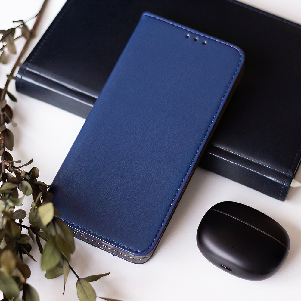 Granatowe etui na telefon leży na białej powierzchni obok czarnego etui na słuchawki i ciemnej książki. Obok widoczne są zielone gałązki roślin.