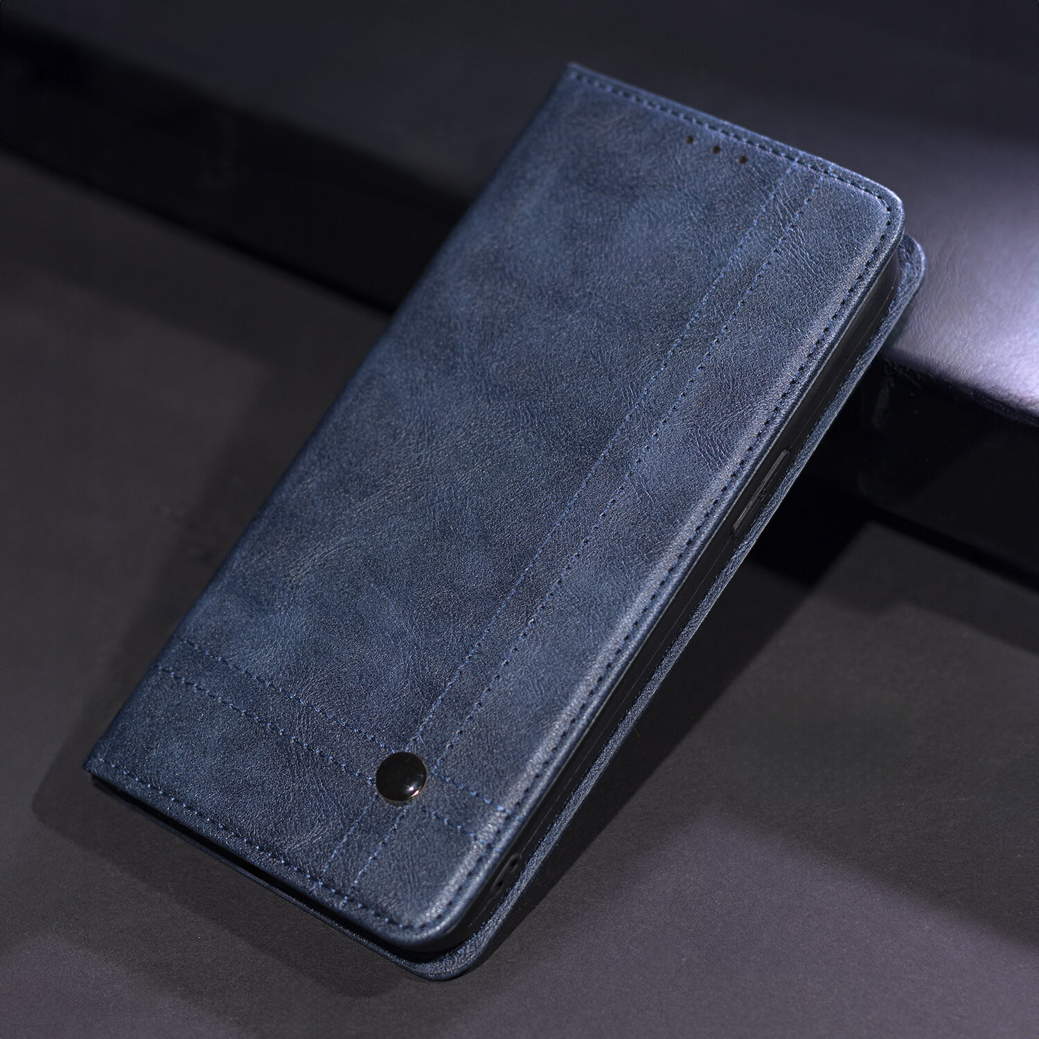 Granatowe etui na telefon z widocznym przeszyciem i metalowym guzikiem na przedniej stronie. Etui leży na ciemnym tle.
