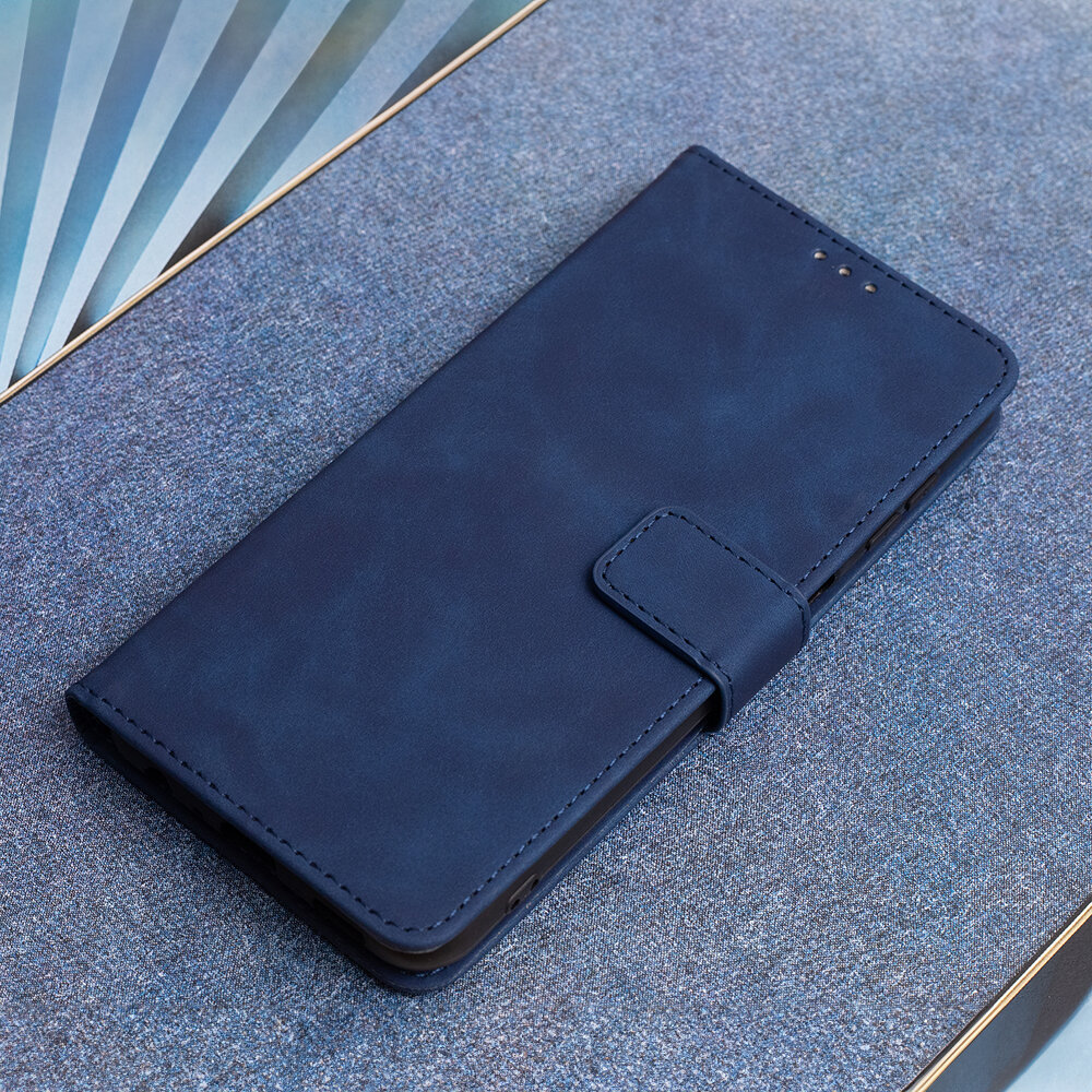 Granatowe etui na telefon z klapką, wykonane z materiału przypominającego welur, leży na powierzchni o podobnym kolorze. Etui posiada zapięcie na magnes i otwory na głośnik.