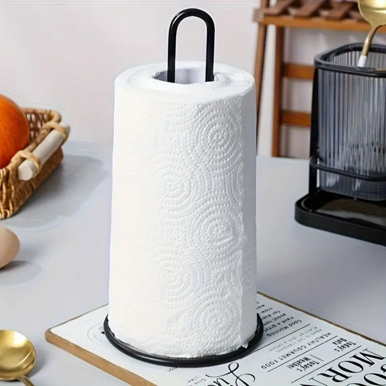 Na obrazie znajduje się stojak na ręcznik papierowy z czarnym metalowym uchwytem, na którym umieszczony jest biały ręcznik papierowy z wzorem w spirale. W tle widoczne są elementy kuchenne, takie jak koszyk z pomarańczami i suszarka na naczynia.