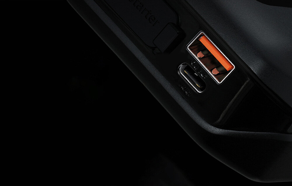 Obraz przedstawia zbliżenie na porty USB urządzenia, w tym jeden port USB-A z pomarańczowym wnętrzem i jeden port USB-C. Tło jest czarne, a powierzchnia urządzenia ma błyszczące wykończenie.
