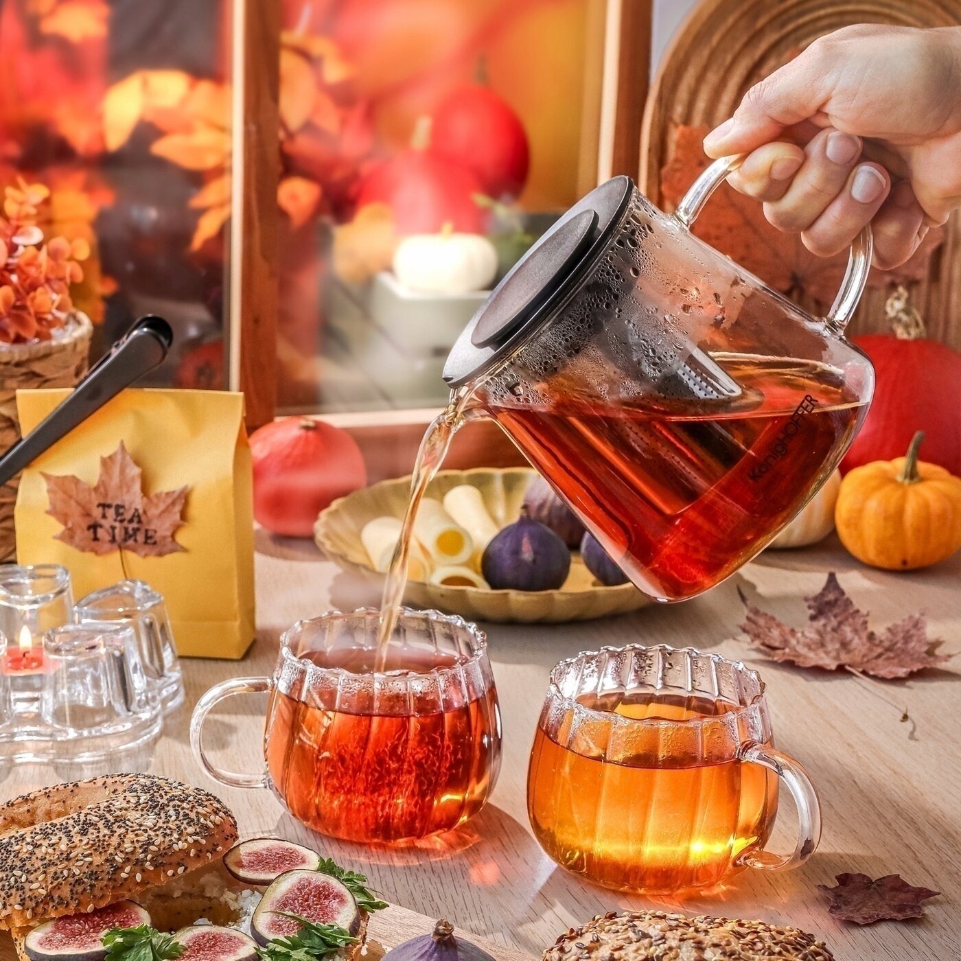 Na stole znajduje się zestaw szklanek z uchem, do których nalewana jest herbata z przezroczystego dzbanka. W tle widoczne są jesienne dekoracje oraz opakowanie z napisem \'TEA TIME\'.
