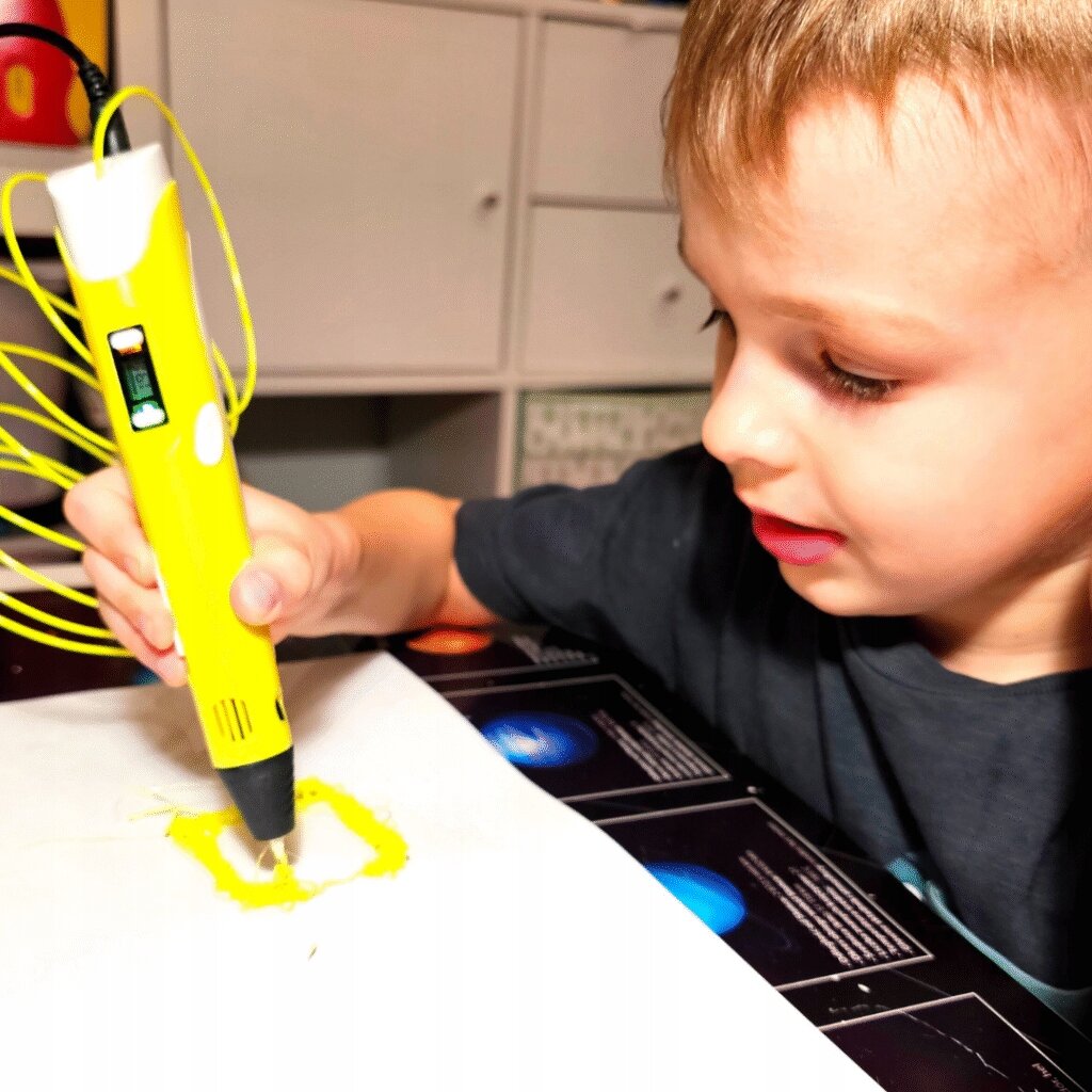 Dziecko używa żółtego długopisu drukarki 3D, tworząc żółty kształt na białej powierzchni. W tle widoczne są półki z szufladami.