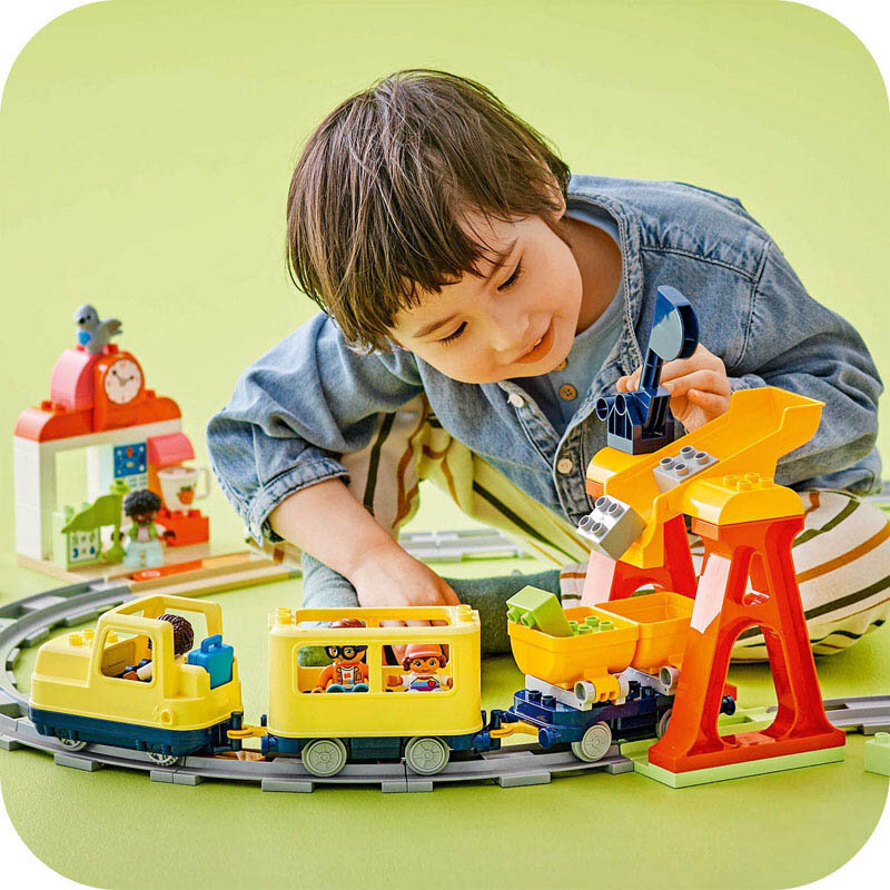 Dziecko bawi się zestawem LEGO DUPLO, w skład którego wchodzi żółty pociąg na torach oraz kolorowy dźwig. W tle widoczna jest budowla z zegarem i figurkami.