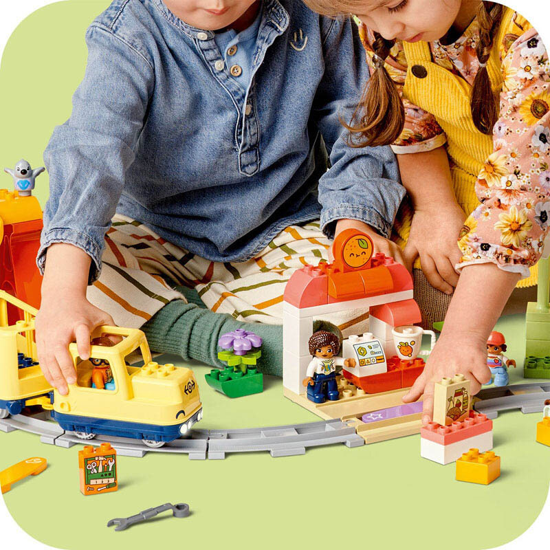 Dwoje dzieci bawi się zestawem LEGO DUPLO, w tym żółtym pociągiem na torach i kolorowymi klockami tworzącymi stację. Na stacji widoczne są figurki, kubek i ekran z rysunkiem misia.