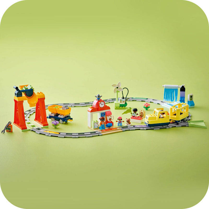 Na zielonym tle znajduje się zestaw LEGO z torami kolejowymi, żółtym pociągiem i różnymi elementami, takimi jak stacja z zegarem i wiatrak. Wokół torów widoczne są figurki i inne kolorowe elementy zestawu.