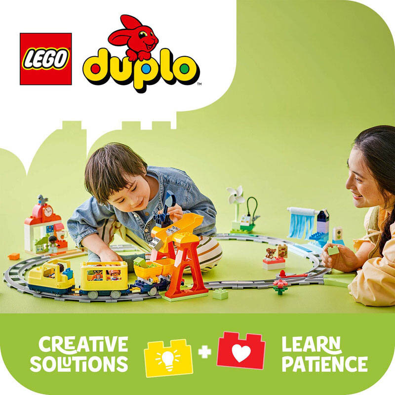 Na obrazku widać dwoje dzieci bawiących się zestawem LEGO DUPLO z pociągiem na torach. W górnej części znajduje się logo LEGO DUPLO, a na dole napis \'CREATIVE SOLUTIONS + LEARN PATIENCE\'.
