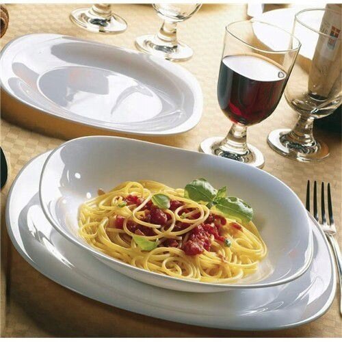 Na stole znajduje się biały talerz z makaronem spaghetti, udekorowany bazylią i sosem pomidorowym. Obok talerza stoi kieliszek z czerwonym winem oraz sztućce.