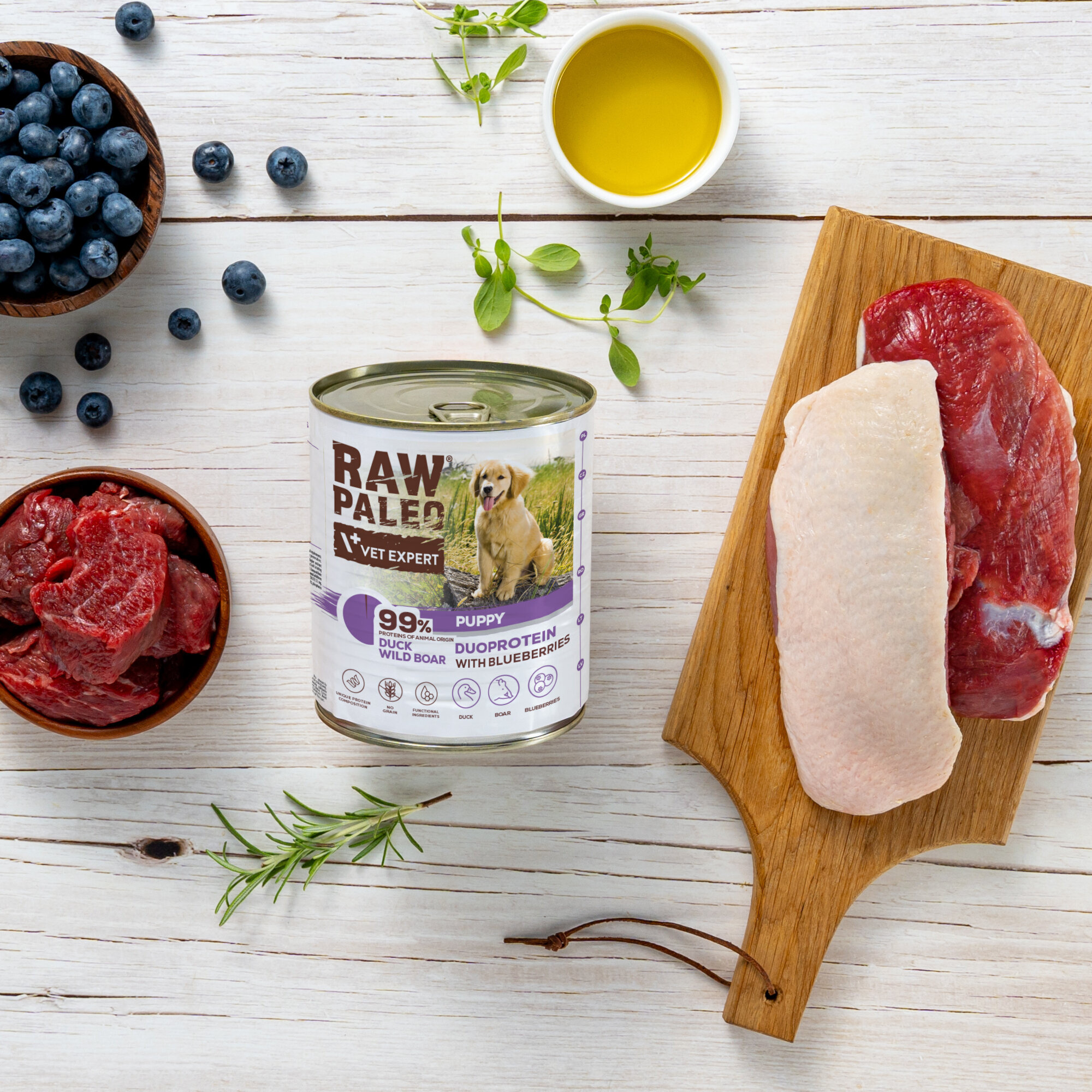 Na drewnianym stole znajduje się puszka karmy RAW PALEO z wizerunkiem psa oraz napisami \'99% DUCK WILD BOAR\' i \'PUPPY DUOPROTEIN WITH BLUEBERRIES\'. Obok puszki leżą kawałki mięsa, miseczka z jagodami, miseczka z surowym mięsem, gałązka rozmarynu i mała miseczka z oliwą.