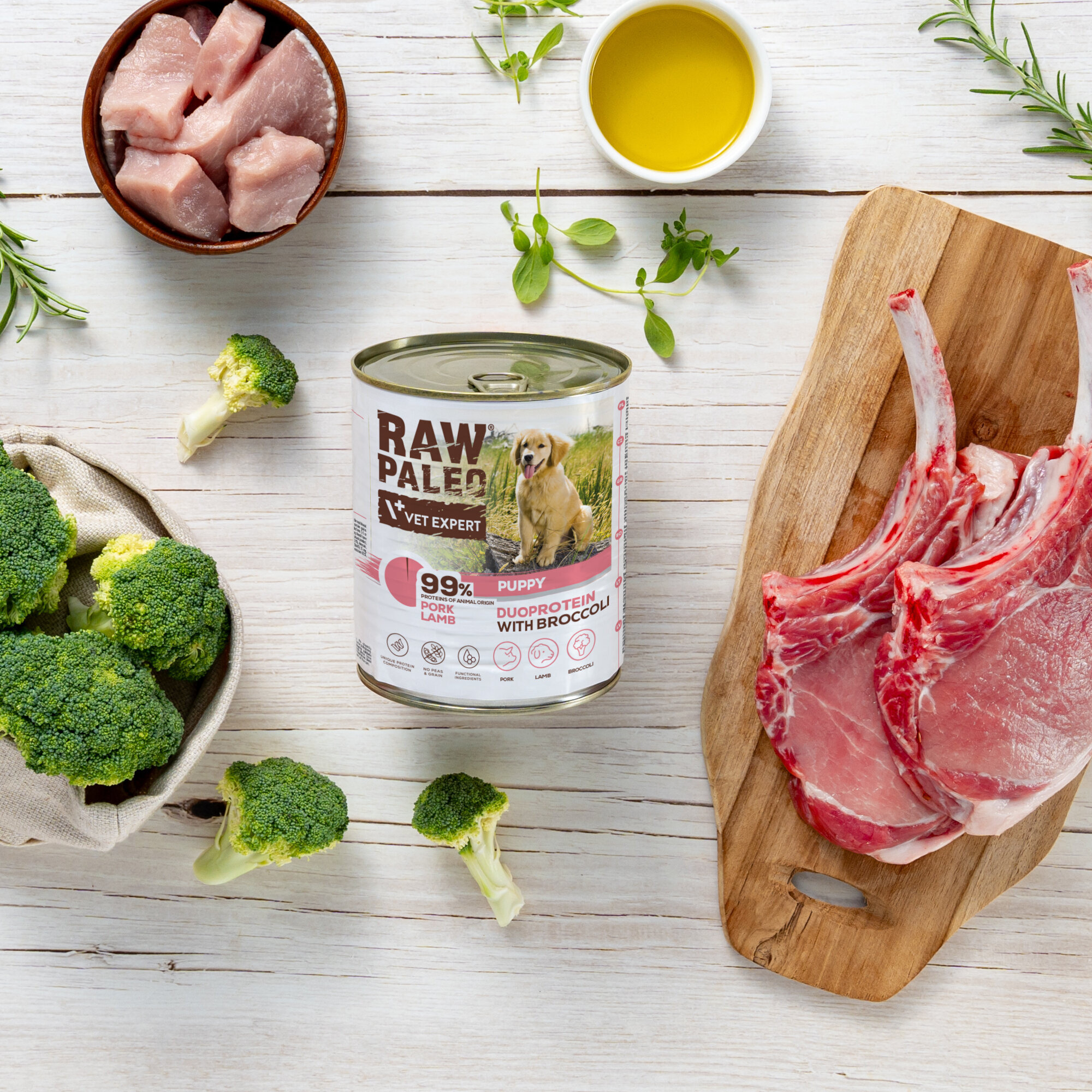 Na drewnianym stole znajduje się puszka karmy dla szczeniąt \'RAW PALEO VET EXPERT\' z wieprzowiną i jagnięciną, otoczona kawałkami mięsa, brokułami, ziołami i miseczką z oliwą.