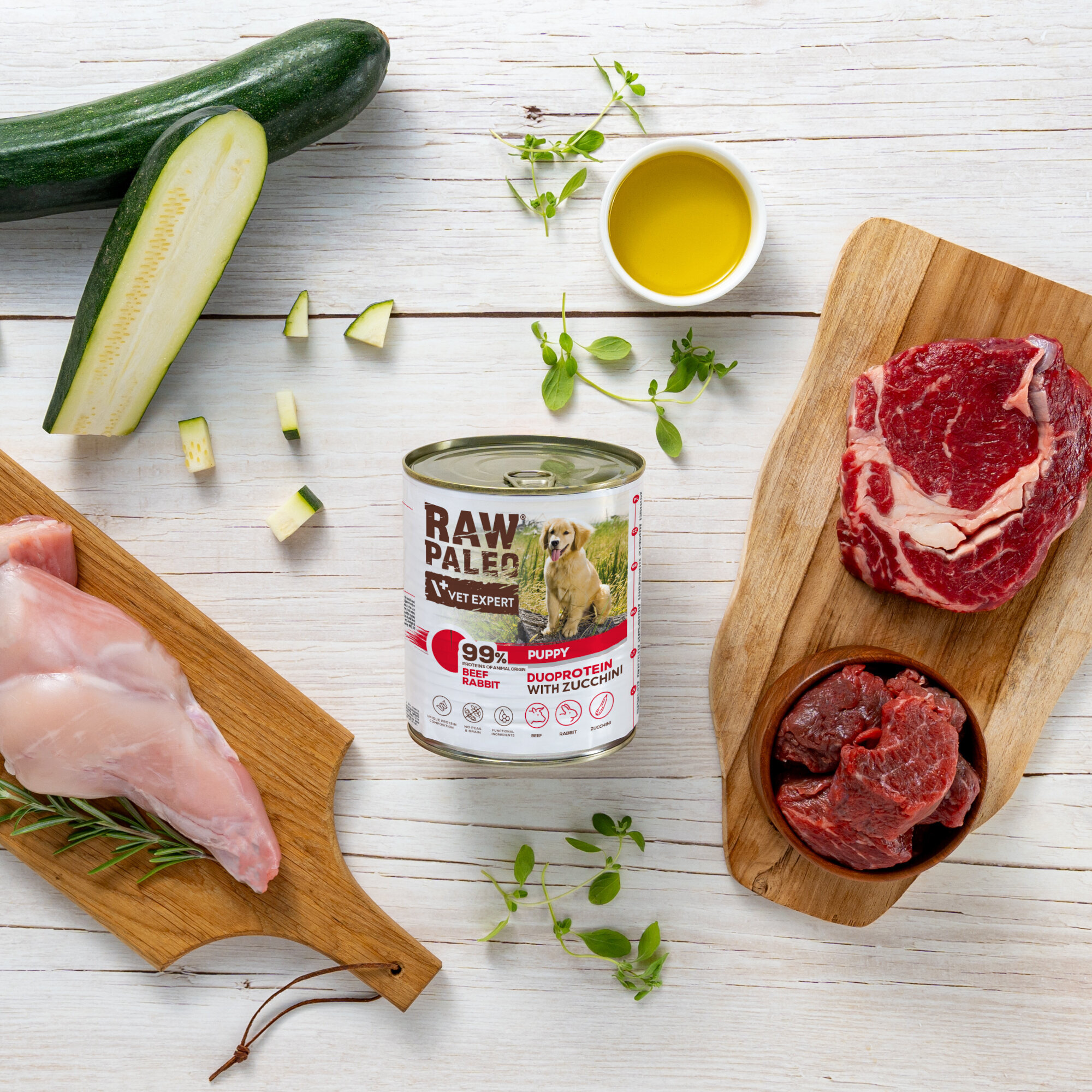 Na drewnianym stole znajduje się puszka karmy RAW PALEO z napisem \'PUPPY DUOPROTEIN WITH ZUCCHINI\', otoczona składnikami takimi jak cukinia, surowe mięso i zioła. Obok puszki leży kawałek surowego mięsa na desce oraz miseczka z oliwą.