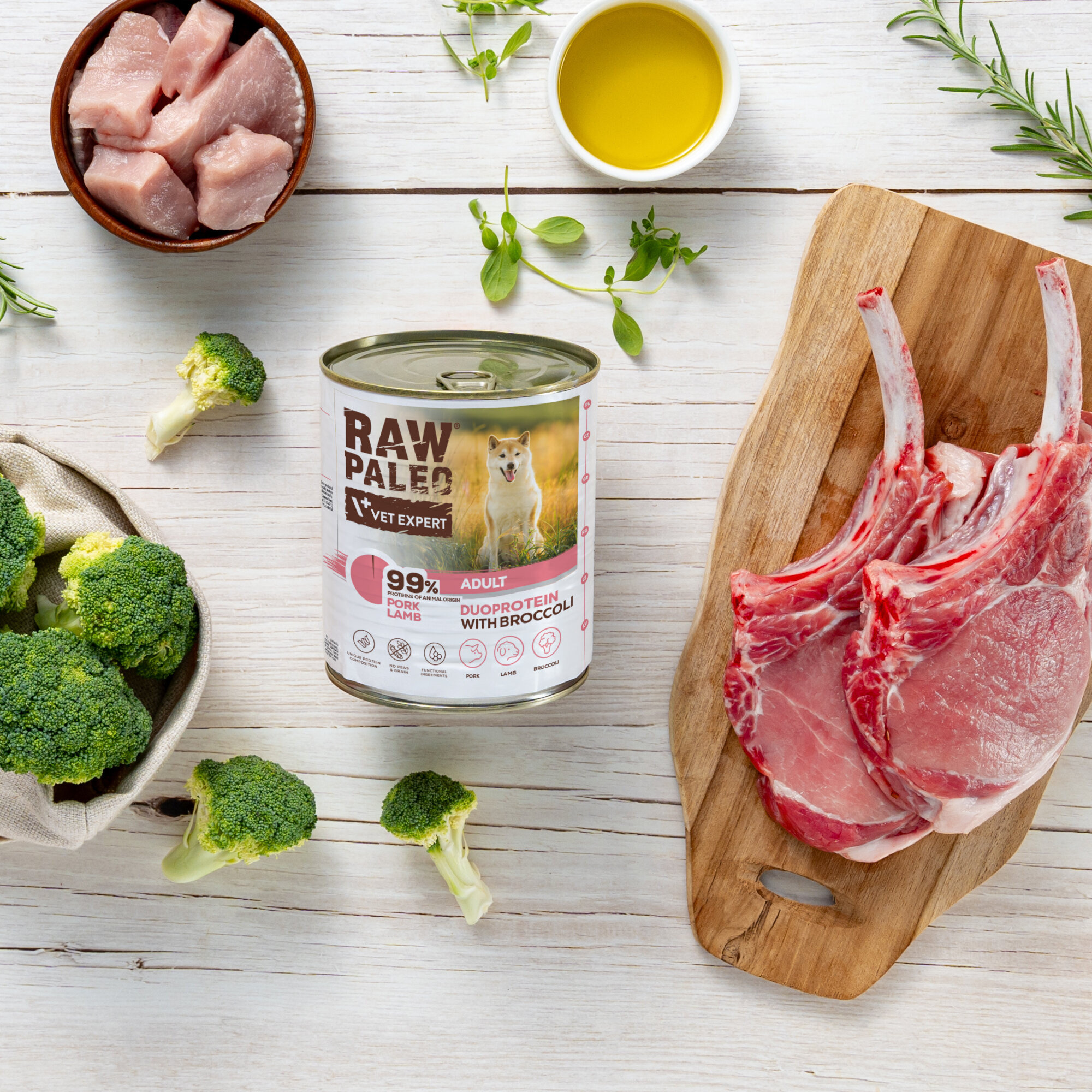 Na drewnianym stole znajduje się puszka karmy dla psów RAW PALEO z napisem \'99% PORK LAMB ADULT DUOPROTEIN WITH BROCCOLI\'. Obok puszki leżą kawałki mięsa, brokuły, miseczka z oliwą oraz zioła.