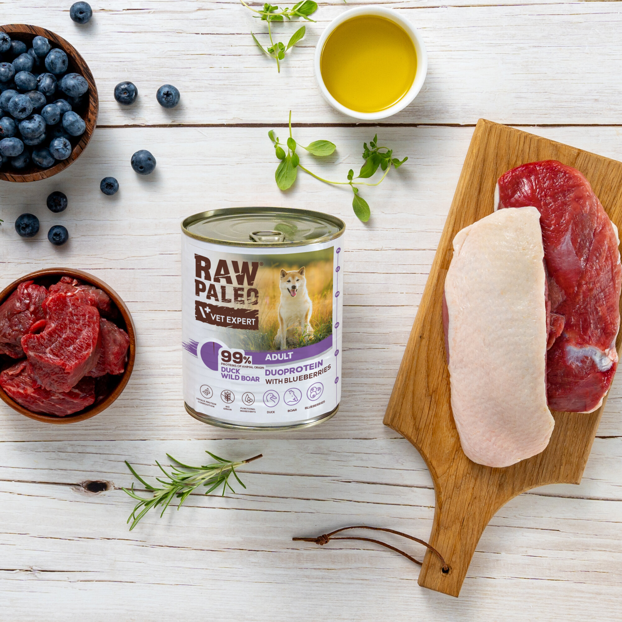 Na drewnianym stole znajduje się puszka karmy dla psów \'RAW PALEO VET EXPERT\' z napisem \'99% DUCK WILD BOAR ADULT DUOPROTEIN WITH BLUEBERRIES\'. Obok puszki leżą kawałki surowego mięsa, jagody, zioła i miseczka z oliwą.