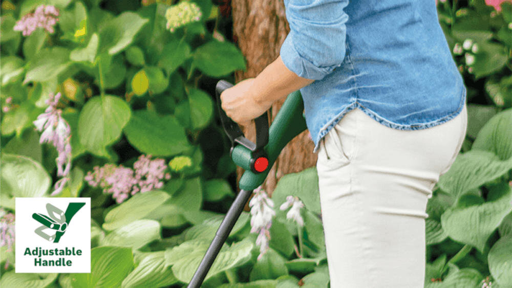 Osoba trzyma podkaszarkę akumulatorową w ogrodzie, otoczona zielonymi roślinami. W lewym dolnym rogu widoczna jest ikona z napisem \'Adjustable Handle\'.