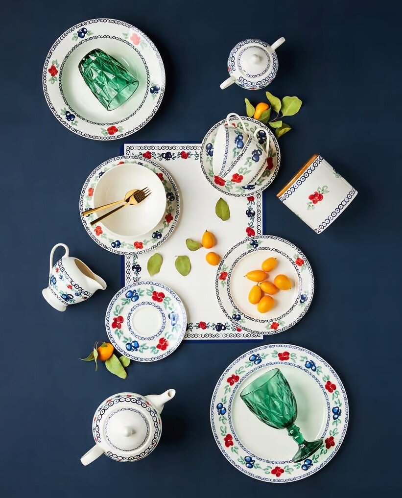 Na ciemnym tle znajduje się zestaw porcelany z motywem kwiatowym, w tym talerze, filiżanki, dzbanek i cukiernica. Na talerzach leżą zielone liście i pomarańczowe owoce, a w jednym z naczyń znajduje się zielony kieliszek.