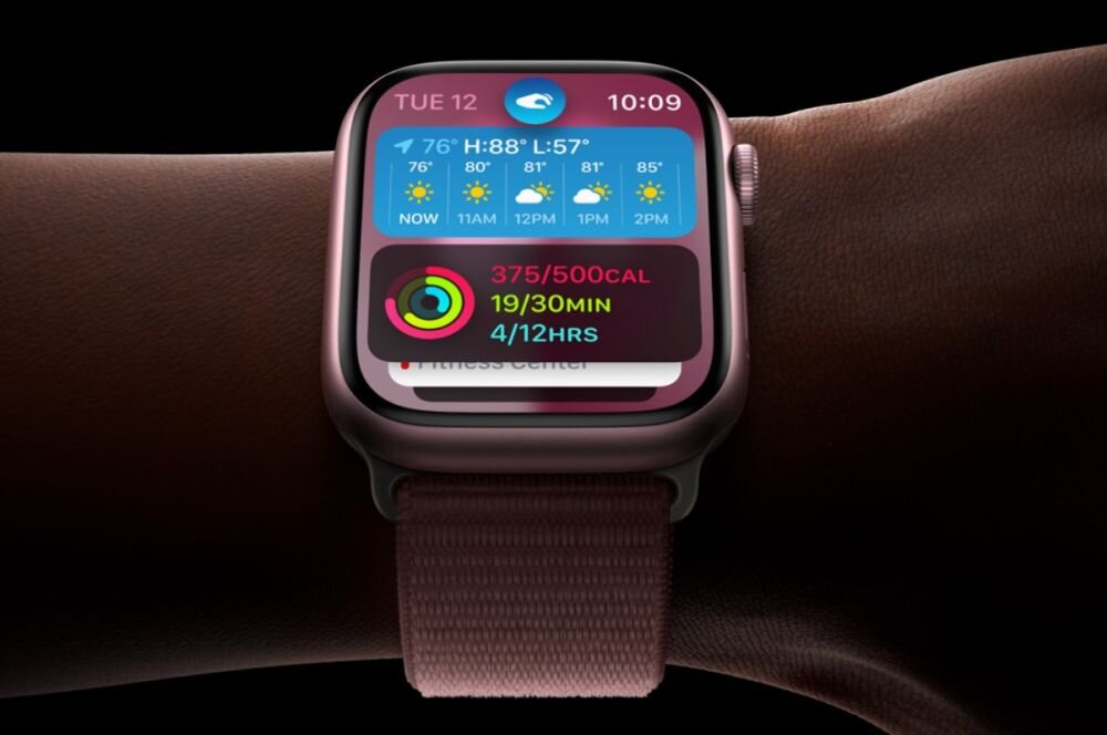 Na zdjęciu widoczny jest smartwatch z wyświetlaczem pokazującym datę, godzinę oraz prognozę pogody. Pasek zegarka jest w kolorze ciemnego błękitu.