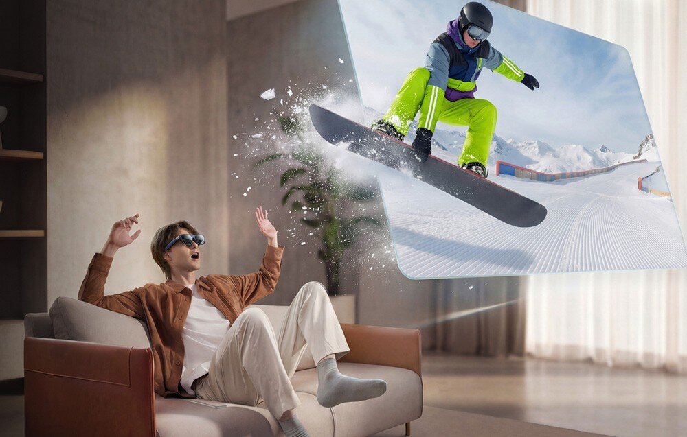 Osoba siedząca na kanapie nosi okulary i patrzy na obraz snowboardzisty unoszącego się w powietrzu, który wydaje się wychodzić z ekranu. W tle widać jasne zasłony i roślinę doniczkową.