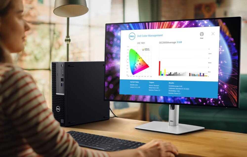 Osoba siedzi przy biurku, patrząc na monitor marki Dell, na którym wyświetlany jest program \'Dell Color Management\' z wykresem i danymi. Obok monitora znajduje się komputer stacjonarny z logo Dell.