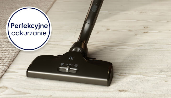 Odkurzacz z logo ELECTROLUX na drewnianej podłodze obok dywanu. Na obrazie widoczny jest napis \'Perfekcyjne odkurzanie\'.