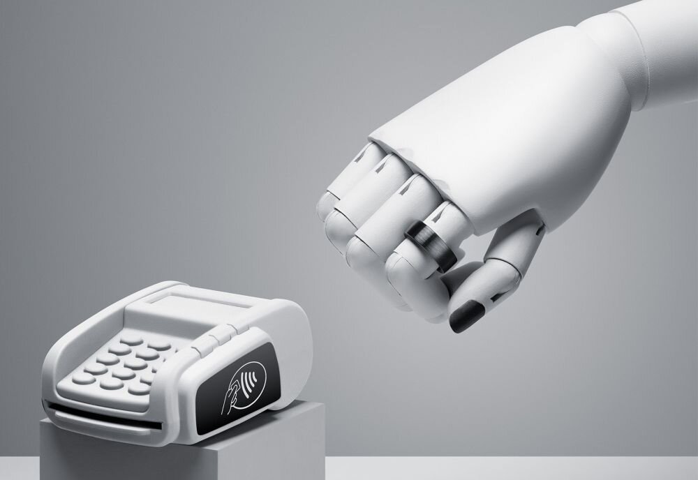 Na obrazie widoczna jest biała, robotyczna ręka zbliżająca się do terminala płatniczego. Terminal ma przyciski i symbol płatności zbliżeniowej na wyświetlaczu.