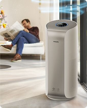 Na pierwszym planie znajduje się oczyszczacz powietrza marki PHILIPS z widocznym panelem sterowania na górze. W tle widać osobę siedzącą na kanapie, czytającą gazetę.