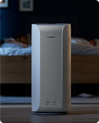 Na zdjęciu znajduje się oczyszczacz powietrza marki PHILIPS w jasnym pomieszczeniu. W tle widoczne jest łóżko z osobami leżącymi pod kołdrą.