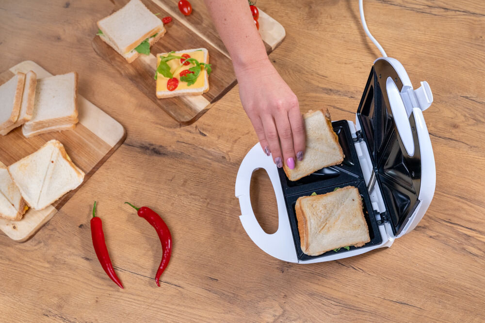 Na drewnianym blacie znajduje się otwarty opiekacz z dwoma tostami, a obok leżą deski z przygotowanymi kanapkami i dwie czerwone papryczki chili. Ręka umieszcza jeden z tostów w opiekaczu.