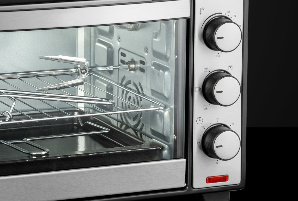 Zdjęcie przedstawia wnętrze mini piekarnika z metalowym rusztem i rożnem. Obok znajdują się trzy pokrętła do regulacji temperatury i funkcji piekarnika.