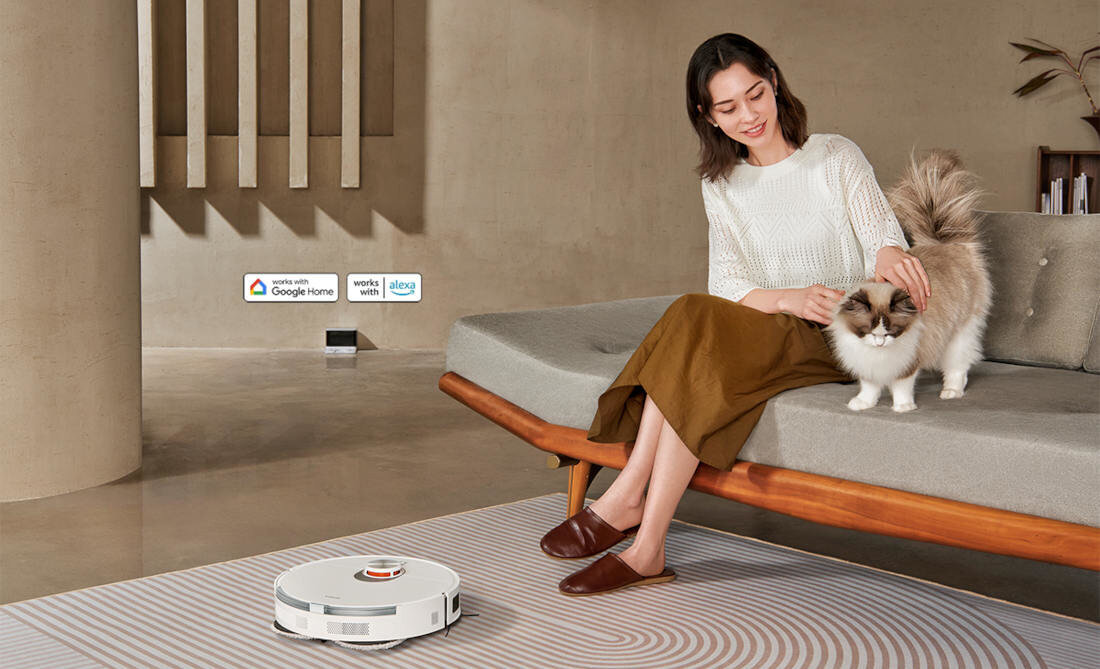 Na obrazie widać biały robot sprzątający na podłodze oraz osobę siedzącą na kanapie, głaszczącą kota. W tle znajdują się loga: \'works with Google Home\', \'works with Alexa\'.