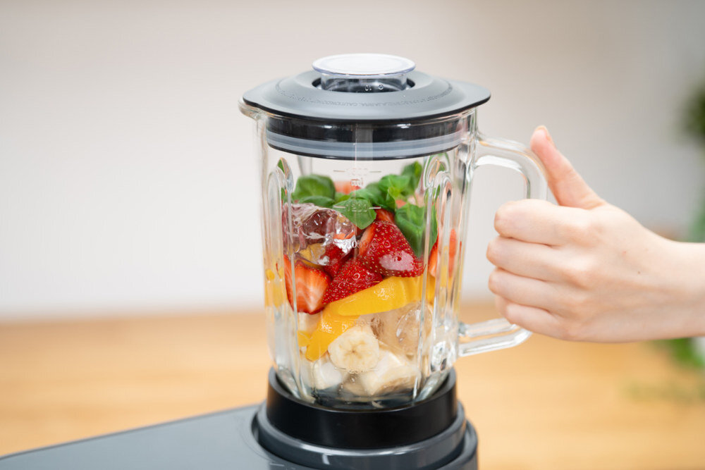 Szklany blender kielichowy wypełniony owocami i liśćmi, trzymany przez rękę. W środku widoczne są truskawki, banany, pomarańcze, zielone liście i kostki lodu.
