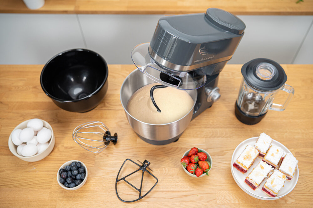 Na drewnianym blacie znajduje się robot kuchenny z misą, w której jest ciasto, obok blender kielichowy, miska z jajkami, miska z jagodami, trzepaczka, mieszadło, miska z truskawkami i talerz z ciastem. Na robocie widoczne jest logo producenta i napis \'media expert\'.