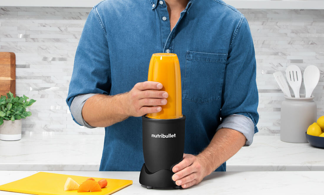 Osoba obsługuje blender personalny z napisem \'nutribullet\' na czarnej podstawie, w której znajduje się żółty pojemnik. Na blacie leżą kawałki owoców i deska do krojenia.