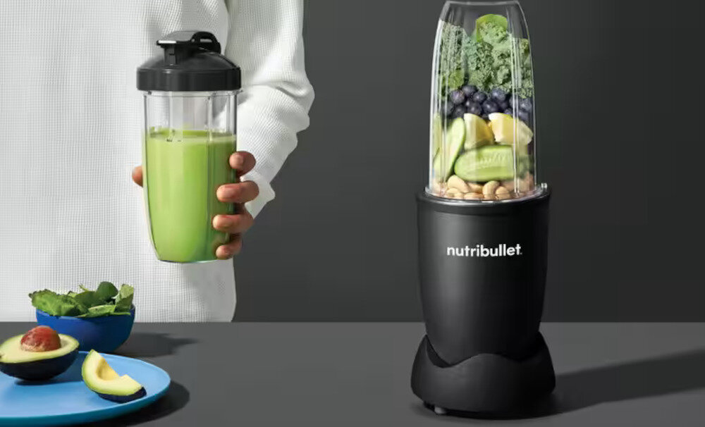 Osoba trzyma kubek z zielonym napojem, obok stoi blender z logo \'nutribullet\' wypełniony owocami i warzywami. Na talerzu leży awokado i liście sałaty.