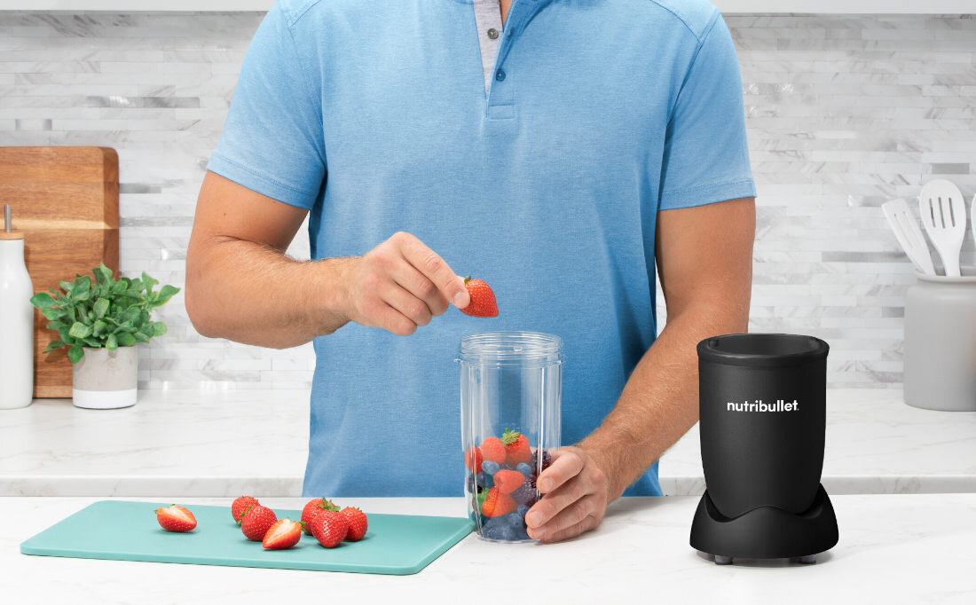 Osoba w niebieskiej koszuli wkłada truskawki do przezroczystego pojemnika blendera stojącego na blacie kuchennym. Obok znajduje się czarny blender z widocznym napisem \'nutribullet\'.
