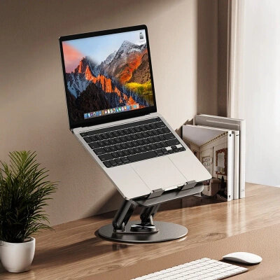 Na biurku znajduje się laptop umieszczony na czarnej, obrotowej podstawce. W tle widoczne są książki i roślina w doniczce.