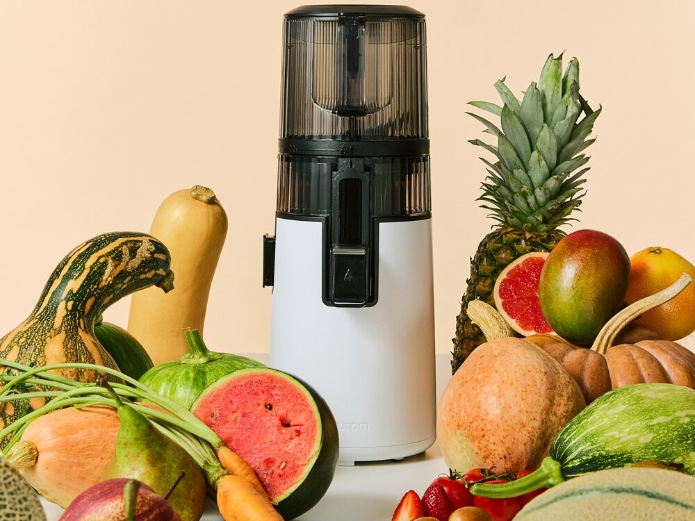 Na obrazie znajduje się wyciskarka wolnoobrotowa w kolorze ciemnoszarym, otoczona różnorodnymi owocami i warzywami, takimi jak ananas, arbuz i dynia. Na urządzeniu widoczne jest logo \'Hurom\'.