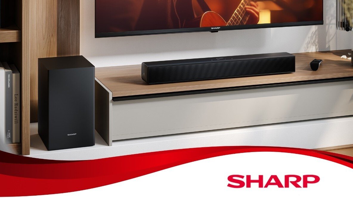 Na obrazie znajduje się czarny soundbar umieszczony na drewnianej półce pod telewizorem oraz czarny subwoofer obok półki. Na dole obrazu widoczne jest czerwone logo z napisem \'SHARP\'.