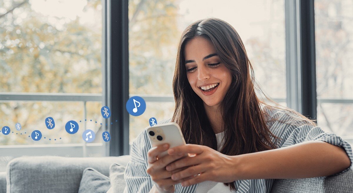 Osoba siedzi na kanapie, trzymając w ręku smartfon. W tle widać ikony Bluetooth i nutki, sugerujące przesyłanie muzyki.