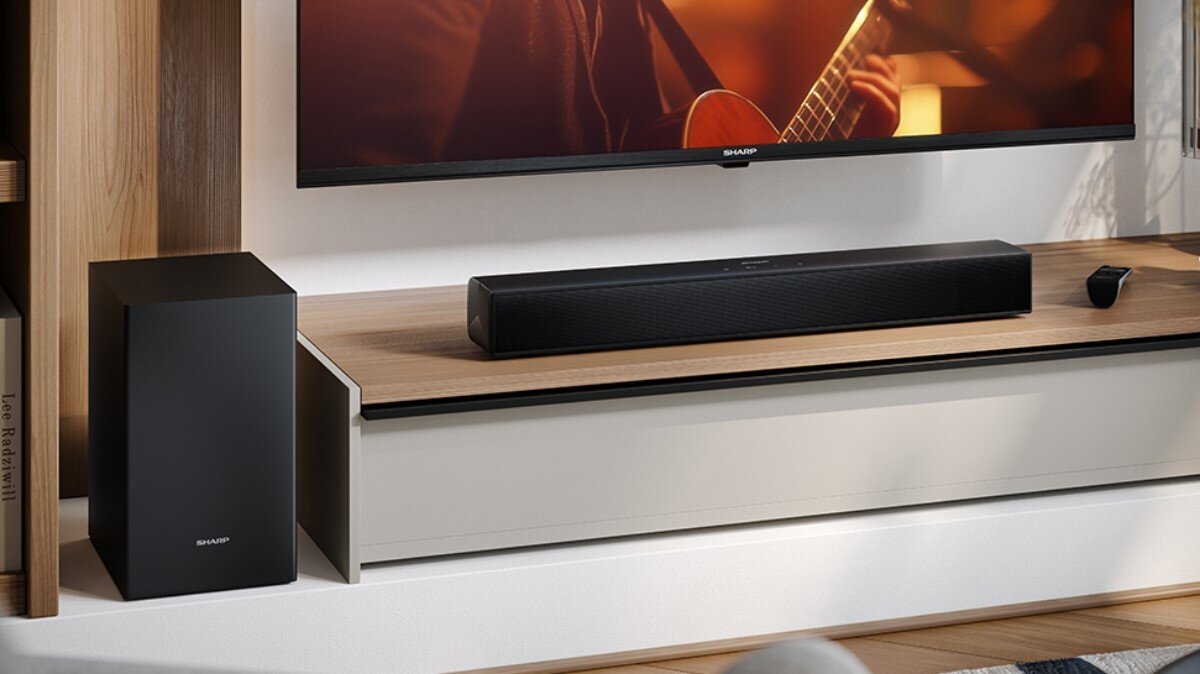 Na obrazie znajduje się czarny soundbar umieszczony na drewnianej półce pod telewizorem, obok którego stoi subwoofer. Na ekranie telewizora widoczna jest scena z osobą grającą na gitarze.