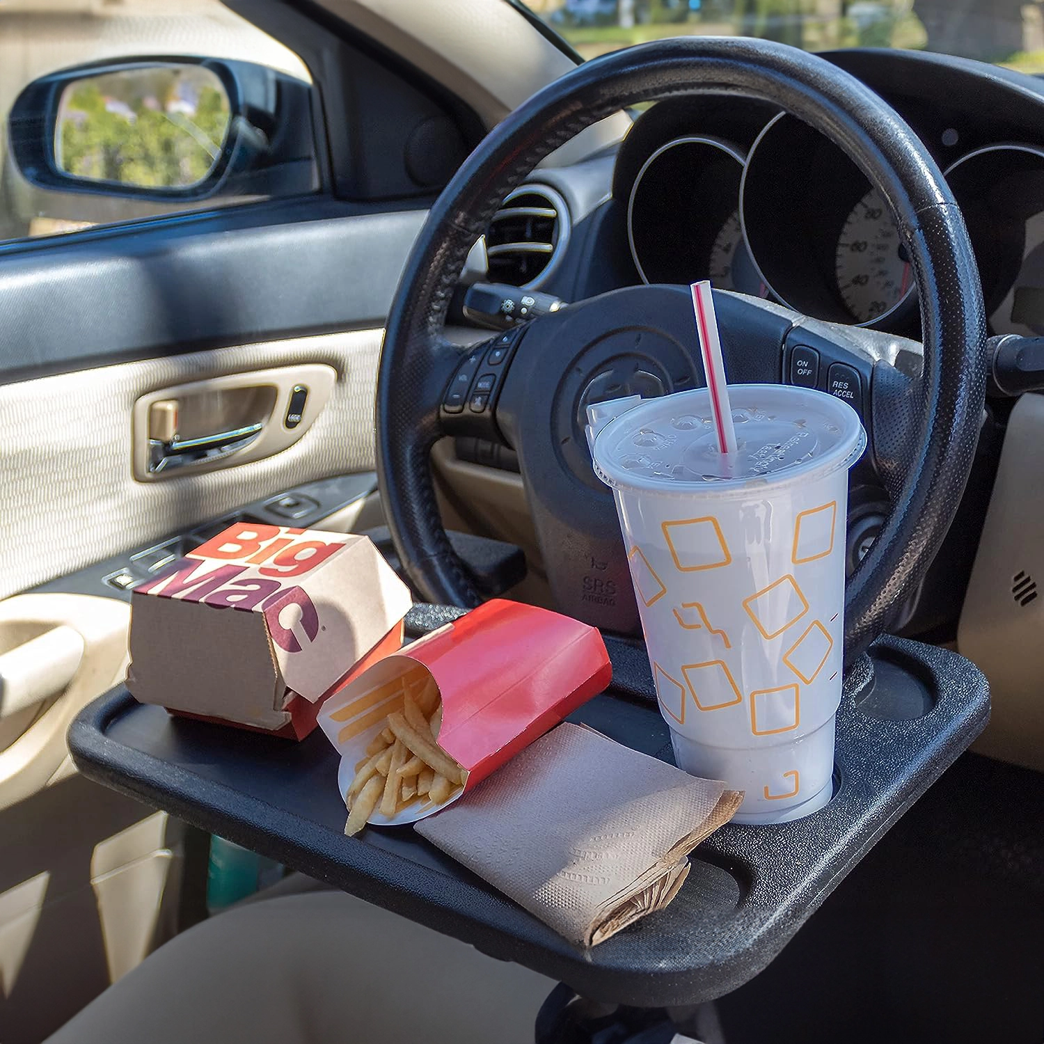 Na zdjęciu widać organizer samochodowy zamontowany na kierownicy, na którym znajduje się kubek z napojem, pudełko z napisem \'Big Mac\', frytki w czerwonym opakowaniu i serwetka. W tle widoczna jest kierownica i wnętrze samochodu.