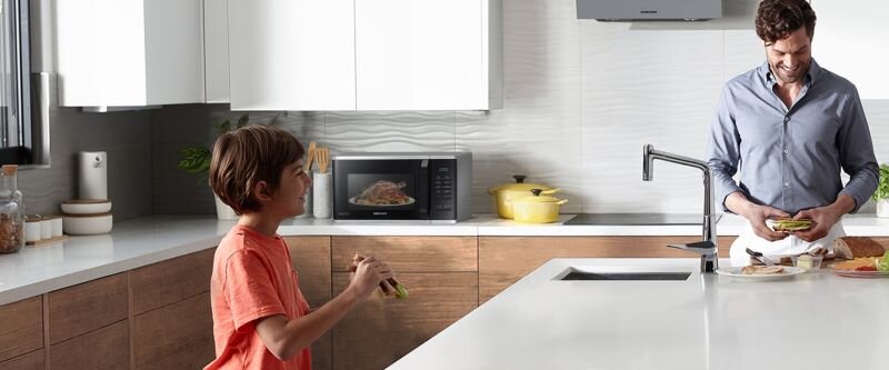 W kuchni znajduje się kuchenka mikrofalowa z widocznym logo SAMSUNG, obok której stoi żółty garnek. Na blacie kuchennym mężczyzna przygotowuje kanapki, a dziecko trzyma jedzenie.